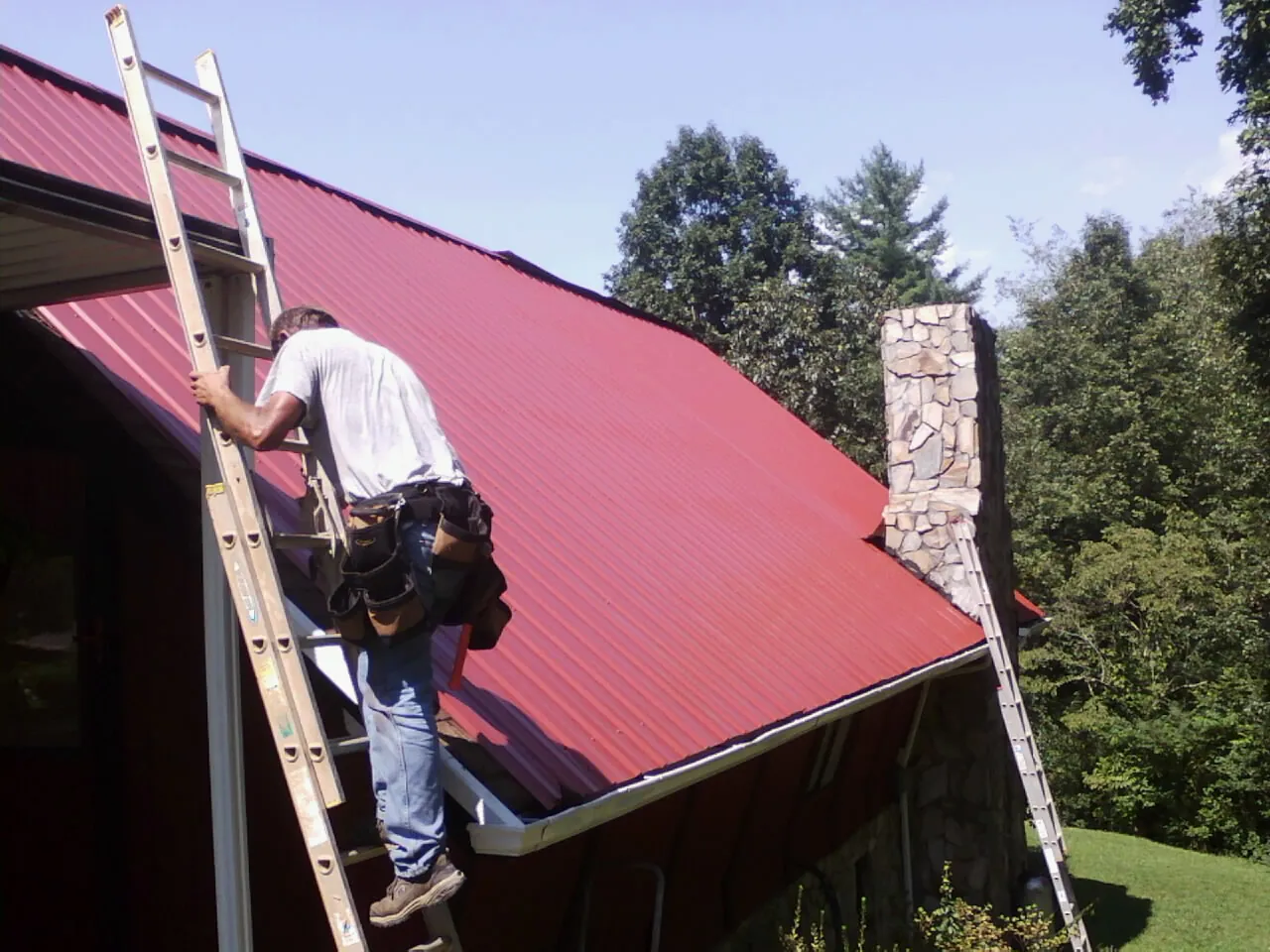 Roofer on a ladder beside a red metal roof and stone chimney — Soffit & Fascia Repair by Roofing Brothers in West Des Moines